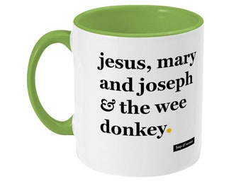 Könnte beinhalten: Weiße Keramiktasse mit grünem Rand und Henkel. Die Tasse hat schwarzen Text, der "jesus, mary and joseph & the wee donkey." lautet. Neben dem Text befindet sich ein kleiner gelber Punkt und unten ein kleines schwarz-weißes Logo mit der Aufschrift "bap & scouse".