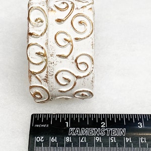 May include: White ceramic cylinder with a gold swirl design. The cylinder is approximately 2 inches in diameter.