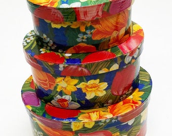 May include: Three round nesting boxes with floral patterns. The boxes are decorated with red, yellow, blue, and green flowers on a dark green background.