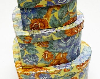May include: Three heart-shaped boxes with a floral pattern. The boxes are stacked on top of each other, with the largest box on the bottom and the smallest box on top. The boxes are made of cardboard and have a yellow background with blue and orange flowers.