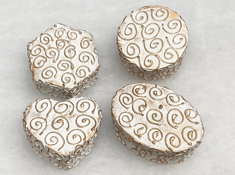 May include: Four white ceramic boxes with a gold swirl design. The boxes are in different shapes, including a heart, a circle, an oval, and a star.