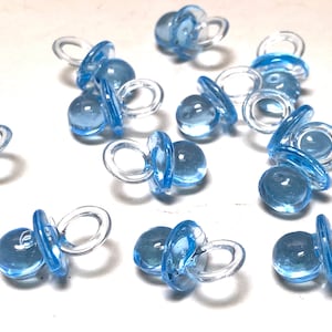 May include: A scattering of light blue plastic baby pacifier charms. The charms are clear with a light blue color and have a small loop for attaching to a gift or party favor.