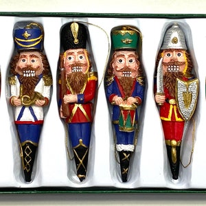 May include: Set of six nutcracker ornaments in a box. The ornaments are made of ceramic and are painted in various colors, including red, blue, green, and gold. Each ornament features a different nutcracker soldier with a unique hat and weapon.