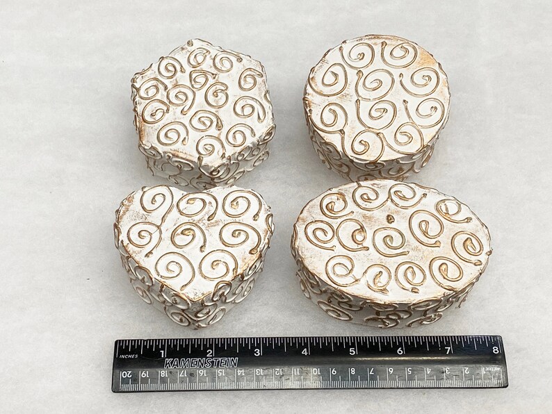 May include: Four white ceramic boxes with gold swirl designs. The boxes are in the shapes of a heart, oval, circle, and hexagon.