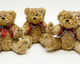 May include: Three plush teddy bears with light brown fur sit side-by-side. Each bear has a red and green plaid bow tie and black button eyes. The bears have a slightly sad expression.
