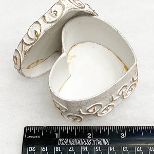 May include: White heart-shaped trinket box with gold swirl design. The box is open and the lid is laying next to it. The box is approximately 2 inches wide.