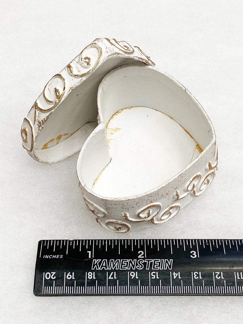 May include: White heart-shaped trinket box with gold swirl design. The box is open and the lid is laying next to it. The box is approximately 2 inches wide.
