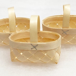 May include: Three small, woven baskets with arched handles. The baskets are made of light-colored wood, with a rectangular shape and a woven pattern. The handles are attached with a small metal X-shaped fastener.