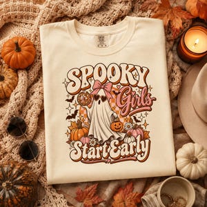 Op de afbeelding: Een crèmekleurig t-shirt met een grafische print van een geest met een roze strik en hartvormige zonnebril, die een discobal en een pompoenemmer vasthoudt. De tekst luidt "SPOOKY Girls Start Early" in een retro, golvend oranje en roze lettertype.