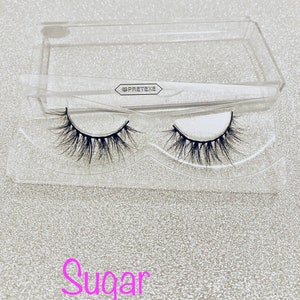 Könnte beinhalten: Ein Paar falsche Wimpern in einem durchsichtigen Plastiketui mit dem Aufdruck "PRETEXTE" auf dem Etui. Die Wimpern sind mit "Sugar" beschriftet.