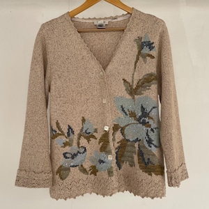 May include: A beige cardigan with a floral design embroidered on the front. The cardigan has a V-neck and a button closure. The floral design is in shades of blue, brown, and green.