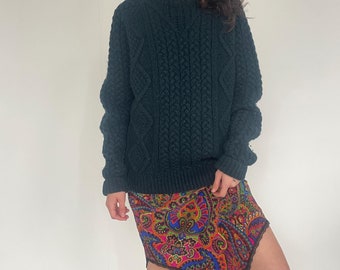 May include: A dark green cable knit sweater paired with a colorful paisley print skirt. The skirt has a unique asymmetrical hemline.