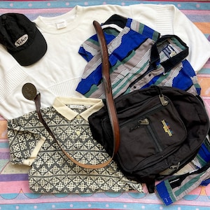 May include: A flat lay of various clothing items, including a white long-sleeved shirt, a black and white patterned polo shirt, a blue and gray striped button-down shirt, a black baseball cap with the word "Nike" on it, and a black backpack with a brown leather strap.