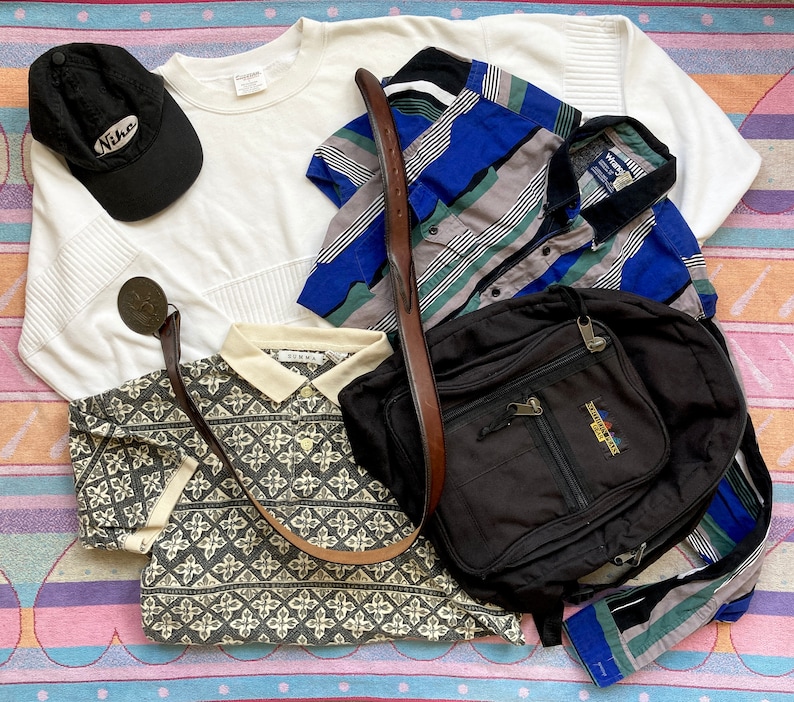 May include: A flat lay of various clothing items, including a white long-sleeved shirt, a black and white patterned polo shirt, a blue and gray striped button-down shirt, a black baseball cap with the word "Nike" on it, and a black backpack with a brown leather strap.