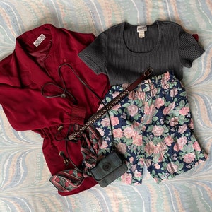 May include: A red jacket with a zipper and a drawstring waist, a gray ribbed knit top, a floral print pair of shorts, and a black and white patterned scarf with a black crossbody bag.