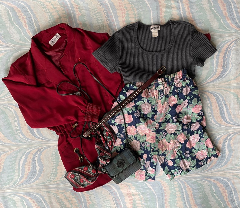 May include: A red jacket with a zipper and a drawstring waist, a gray ribbed knit top, a floral print pair of shorts, and a black and white patterned scarf with a black crossbody bag.
