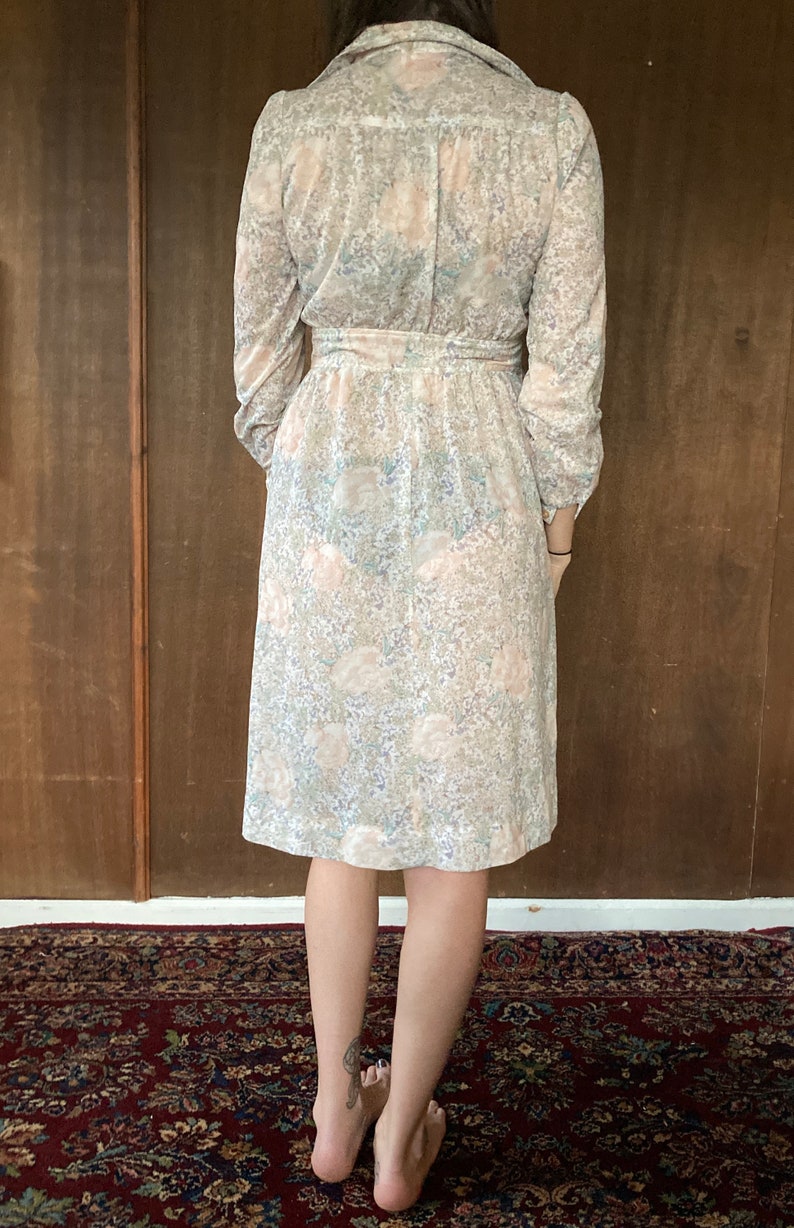 May include: A long-sleeved dress with a floral print in shades of pink, white, and blue. The dress has a collared neckline, a belt at the waist, and a knee-length skirt.