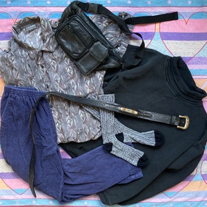 May include: A flat lay of clothing items including a blue corduroy pants, a black sweatshirt, a gray and brown patterned shirt, a black leather fanny pack, and a brown leather belt.