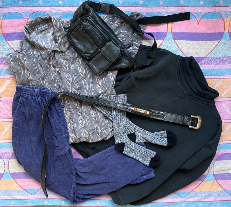 May include: A flat lay of clothing items including a blue corduroy pants, a black sweatshirt, a gray and brown patterned shirt, a black leather fanny pack, and a brown leather belt.