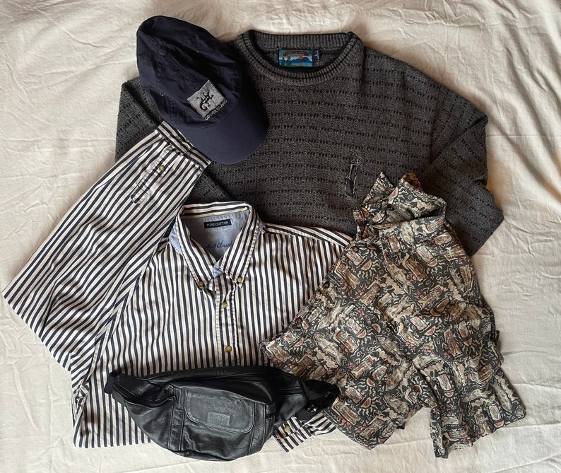 May include: A flat lay of a variety of clothing items, including a striped button-down shirt, a gray sweater, a floral print shirt, a black fanny pack, and a blue baseball cap with a logo.