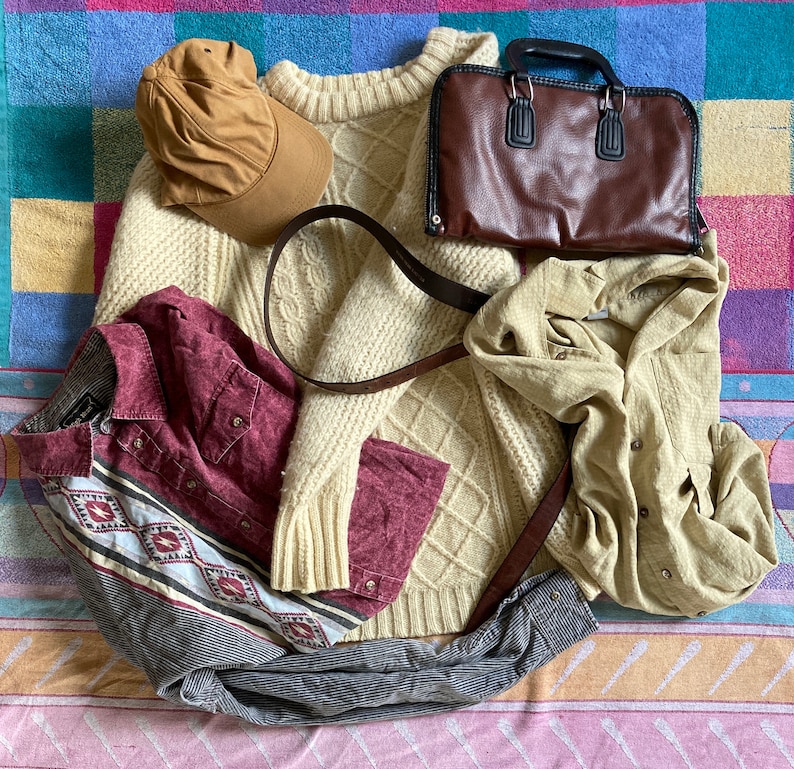 May include: A flat lay of a beige cable knit sweater, a brown leather bag, a brown felt hat, a beige and brown plaid shirt, and a beige linen shirt. The items are arranged on a blue and pink patterned fabric.