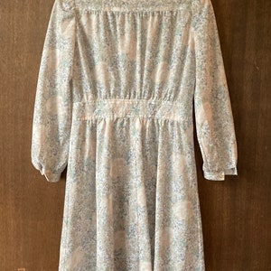 May include: A white dress with a floral print. The dress has a collared neckline, long sleeves, and a fitted waist. The dress is hanging on a wooden hanger.