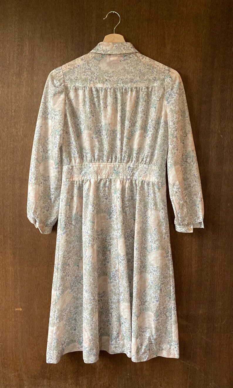 May include: A white dress with a floral print. The dress has a collared neckline, long sleeves, and a fitted waist. The dress is hanging on a wooden hanger.