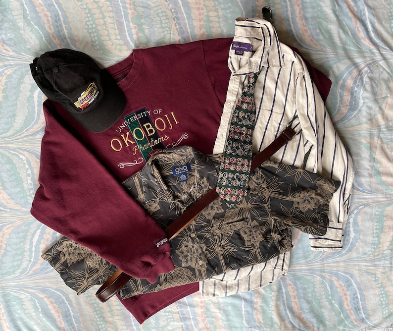 May include: A maroon sweatshirt with the text "University of Okoboji Phantoms" in white and yellow embroidery. A black and white patterned shirt with a brown leather belt is layered over the sweatshirt. A black baseball cap with the text "Exceptional" in white is on top of the sweatshirt. A white and blue striped button-down shirt with a patterned tie is on top of the other shirts.