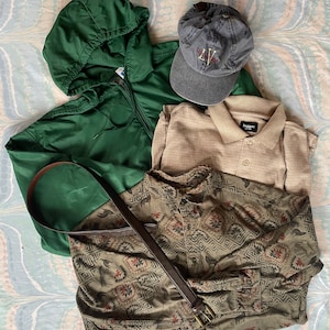 May include: A flat lay of men's clothing items including a green hooded jacket, a beige polo shirt, a green and brown patterned button-down shirt, a brown leather belt, and a blue baseball cap with the text "Los Vegas" embroidered on the front.