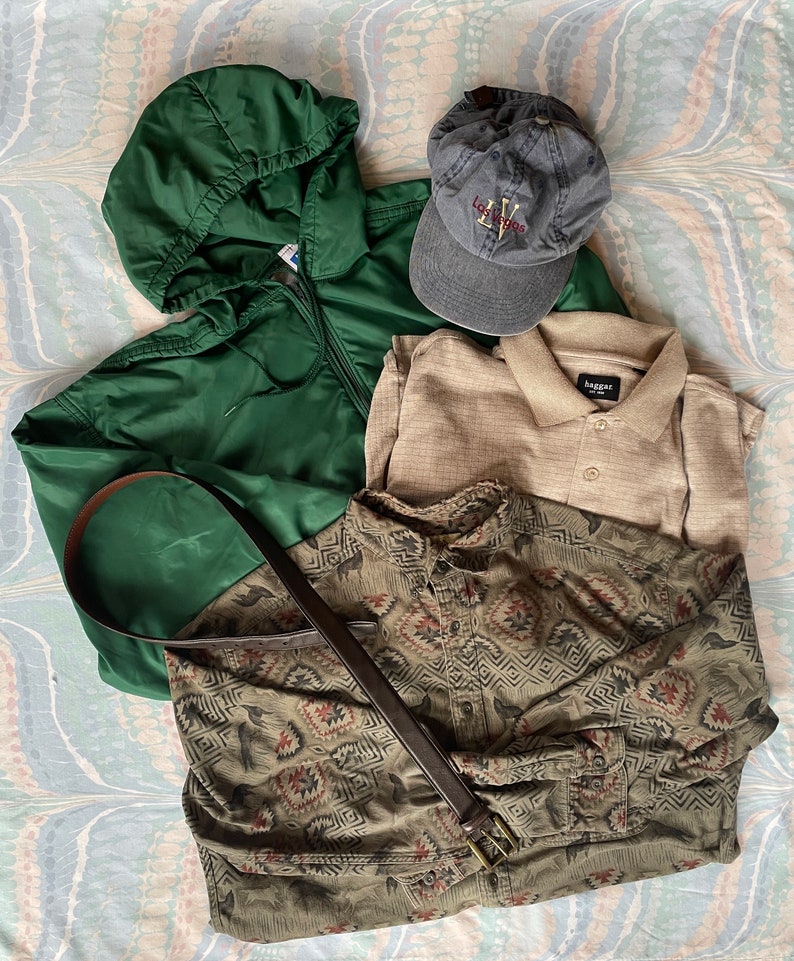May include: A flat lay of men's clothing items including a green hooded jacket, a beige polo shirt, a green and brown patterned button-down shirt, a brown leather belt, and a blue baseball cap with the text "Los Vegas" embroidered on the front.