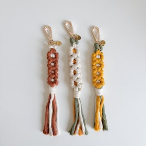 May include: Three handmade macrame keychains with a tassel. The keychains are made with brown, green, and yellow string and feature a daisy flower design.