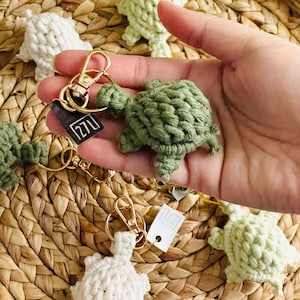 May include: A set of five crocheted turtle keychains in various shades of green and white. The keychains are attached to gold key rings and feature a small brown tag with the text "1211".