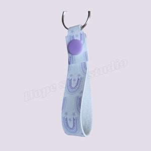 May include: White leather keychain with a purple button and a purple and white pattern of rainbows and stars.