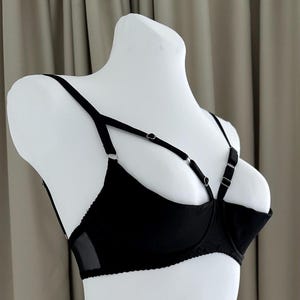 May include: Black bra with adjustable straps and a unique design. The bra features a sheer mesh panel on the side and a decorative strap design. The bra is displayed on a white mannequin.
