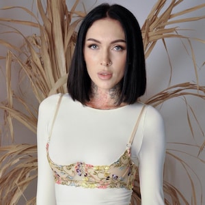 May include: A cream-colored long-sleeve top with a floral embroidered bra. The bra features a beige base with colorful floral embroidery in shades of pink, purple, yellow, and green. The top has thin straps and a scoop neckline.