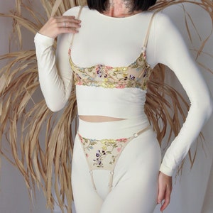Könnte beinhalten: Cremefarbenes Dessous-Set mit einem langärmeligen Oberteil, einem floral bestickten BH und einem passenden String. Der BH und der String sind mit farbenfrohen Blumenstickereien verziert. Das Set ist aus weichem, bequemem Material.