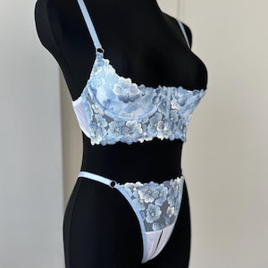 Könnte beinhalten: Hellblaues Dessous-Set mit floralen Spitzendetails. Das Set beinhaltet einen BH mit verstellbaren Trägern und einen passenden String. BH und String sind mit zarten, gestickten Blumen verziert, was einen romantischen Look erzeugt.