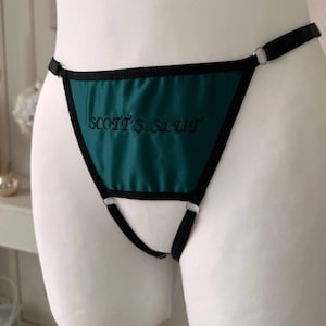 May include: Teal thong underwear with black trim and the embroidered text "SCOTT'S SLUT". The lingerie features a triangle front panel and thin straps. A matching teal bra with black trim is visible in the background.