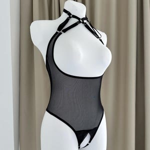 Puede incluir: Body de malla negra con cuello halter y diseño de entrepierna abierta. La lencería presenta correas ajustables y detalles de anillos metálicos. La prenda se exhibe en un maniquí blanco sobre un fondo neutro.