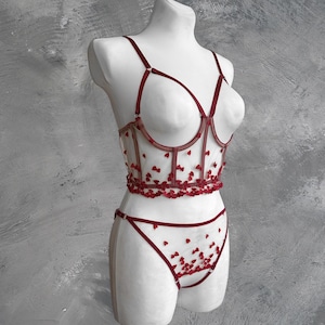 Puede incluir: Un conjunto de lencería delicado con tela de encaje roja transparente y un diseño en forma de corazón. El conjunto incluye un sujetador y una braga.