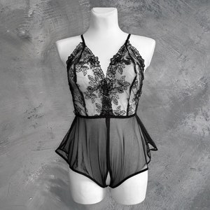 Op de afbeelding: Zwart lingerie body met bloemen kanten details. De doorschijnende stof heeft een V-hals en een delicaat ontwerp met open kruis. De body wordt getoond op een witte mannequin tegen een grijze achtergrond.