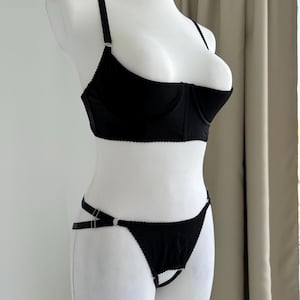Peut inclure: Ensemble de lingerie noir composé d'un soutien-gorge et d'un string. Le soutien-gorge est doté d'armatures, de bretelles réglables et d'un bord festonné. Le string présente un design de bretelles unique avec des anneaux métalliques. L'ensemble est présenté sur un mannequin blanc.