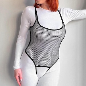 May include: Black fishnet bodysuit with thin straps over a white long-sleeve top and white leggings. The bodysuit has a scoop neckline and a high-cut leg design. The outfit is form-fitting and has a modern, edgy aesthetic.