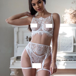 Op de afbeelding: Witte kanten lingerie set met een beha, een slip met hoge taille en een jarretellegordel. Het delicate bloemen kanten ontwerp bedekt de bh-cups en de voorkant van de slip. Verstelbare bandjes en jarretellegordels maken de set compleet. Op de achtergrond staat een witte piano.