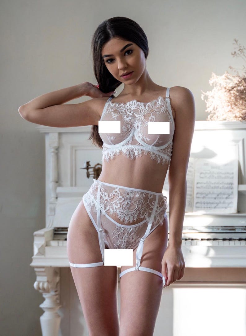 Op de afbeelding: Witte kanten lingerie set met een beha, een slip met hoge taille en een jarretellegordel. Het delicate bloemen kanten ontwerp bedekt de bh-cups en de voorkant van de slip. Verstelbare bandjes en jarretellegordels maken de set compleet. Op de achtergrond staat een witte piano.