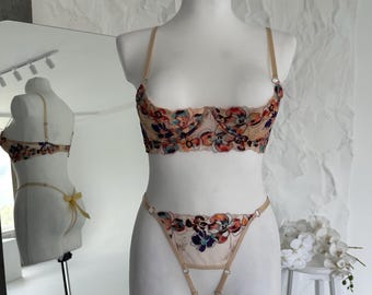 May include: A beige lingerie set with floral embroidery in red, blue, and orange. The set includes a bra and a thong, displayed on a mannequin. The bra has adjustable straps. The background is a white wall and a mirror.