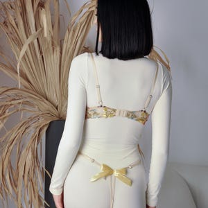 May include: A white long-sleeved top with a floral lace bra and a yellow ribbon detail. The top is worn with white trousers.