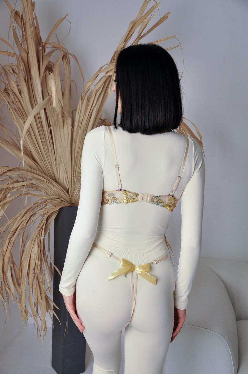 May include: A white long-sleeved top with a floral lace bra and a yellow ribbon detail. The top is worn with white trousers.
