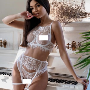 Op de afbeelding: Witte kanten lingerie set met een beha, een slip met hoge taille en jarretels. Het delicate bloemen kanten ontwerp is zichtbaar. De set wordt getoond op een persoon die tegen een witte piano leunt.
