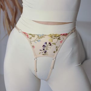May include: White lingerie set featuring floral embroidery. The top is a cropped tank top with a delicate lace trim. The bottom is a garter belt with a floral design and adjustable straps.